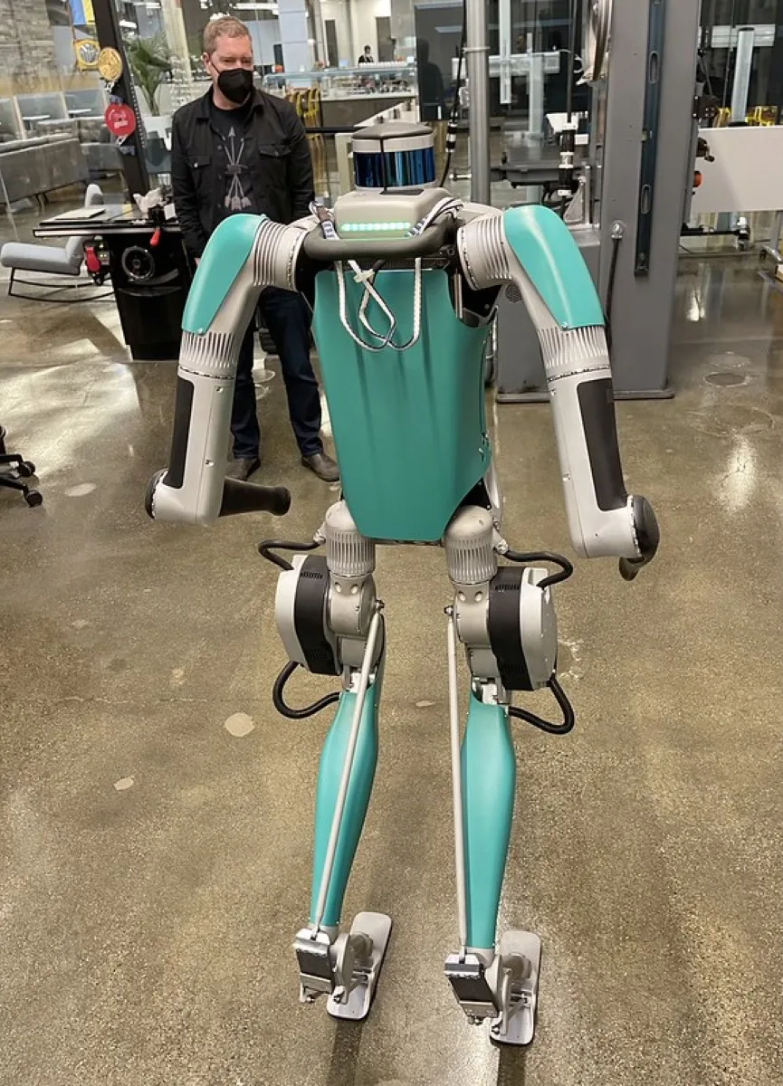Agility Robotics Digit, a teal and grey bipedal robot with reverse-knee legs, standing indoors