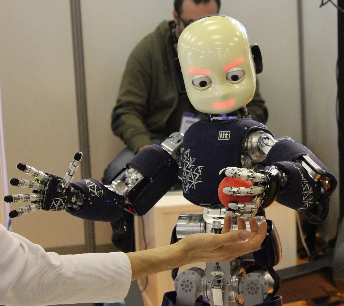 iCub humanoid research robot reaching toward a person's hand holding an orange ball, at the Innorobo 2014 exhibition in Lyon