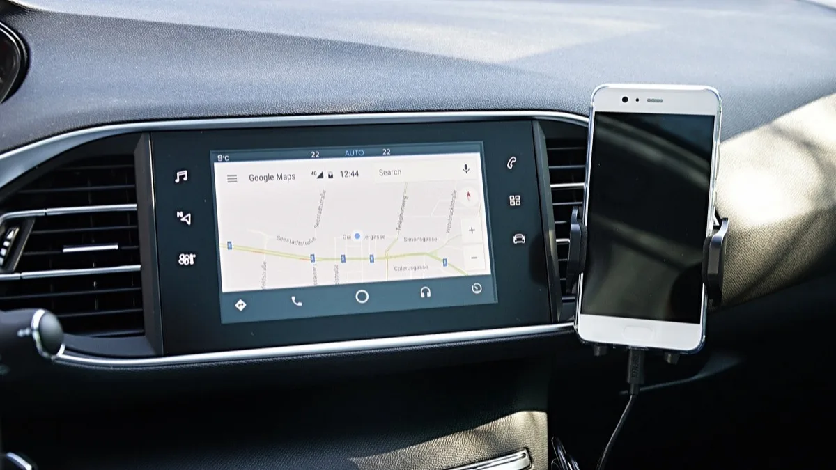 A phone mounted on a car dashboard and wired into an Android Auto head unit showing Google Maps