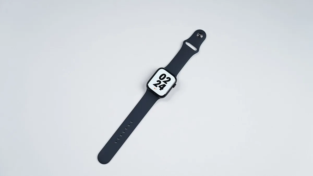 An Apple Watch with a dark sport band and a square face lying flat on a white surface