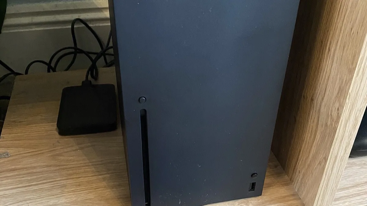 A black Xbox Series X console standing upright on a wooden shelf beside a cable and power brick