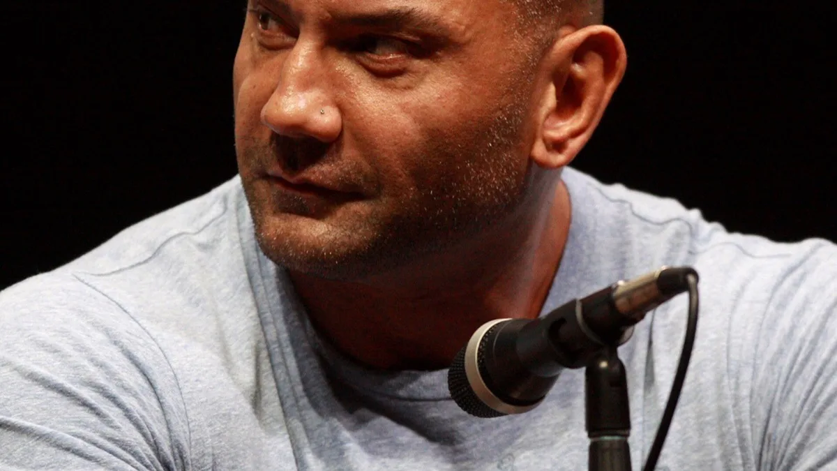 Dave Bautista speaking into a microphone at a convention panel against a dark backdrop