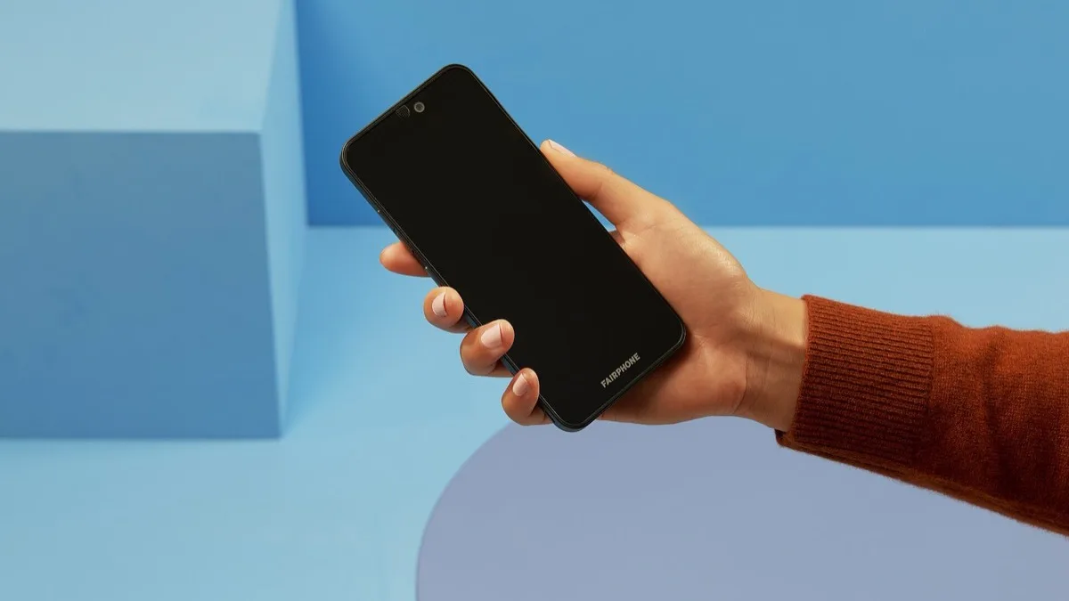 A hand holding the Fairphone in front of a blue backdrop, showing the phone's front camera cutout and Fairphone branding on the chin