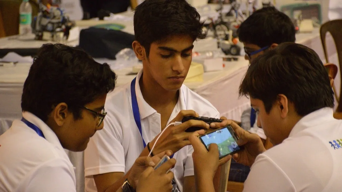 Four students crowded around a smartphone held sideways in landscape mode, watching a game running on its screen
