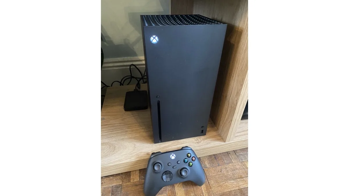 A black Xbox Series X console standing on a wooden shelf with its matching wireless controller in front of it
