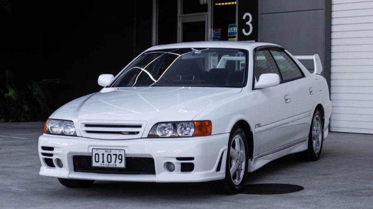 A white Toyota Chaser Tourer V JZX100 in TRD Sports trim parked outside a garage, one of the cars added in Gran Turismo 7 update 1.71