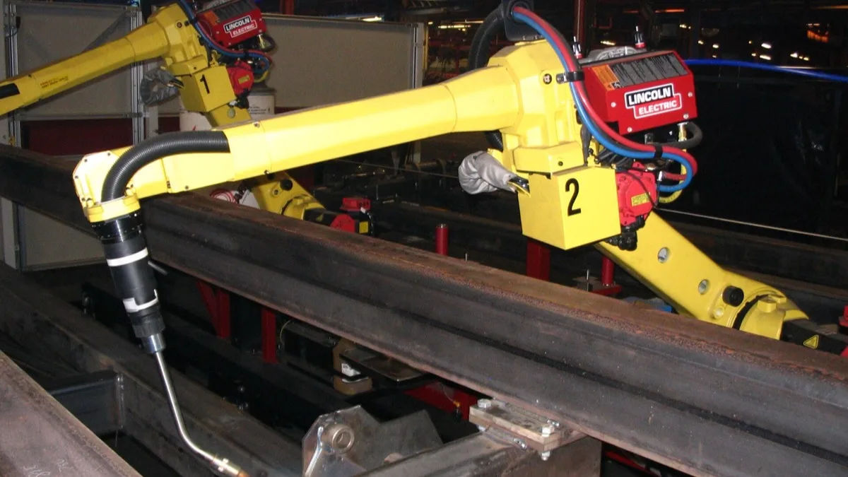 A yellow FANUC industrial welding robot arm fitted with a Lincoln Electric welding head, working on a steel beam on a factory line