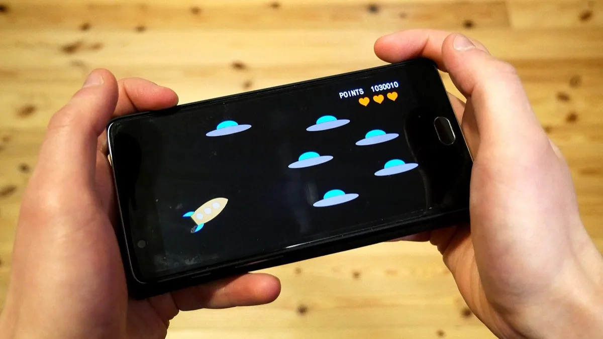 Two hands holding an Android phone in landscape mode, playing a simple space shooter game above a wooden table