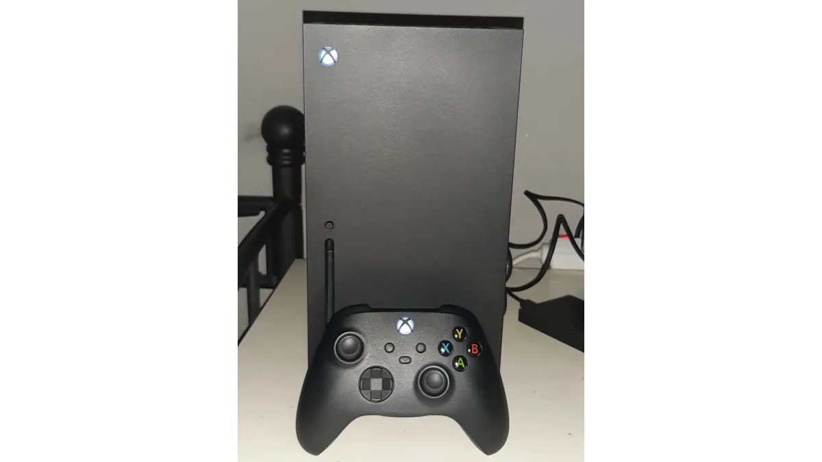 An Xbox Series X console standing upright on a desk with a black Xbox wireless controller resting in front of it