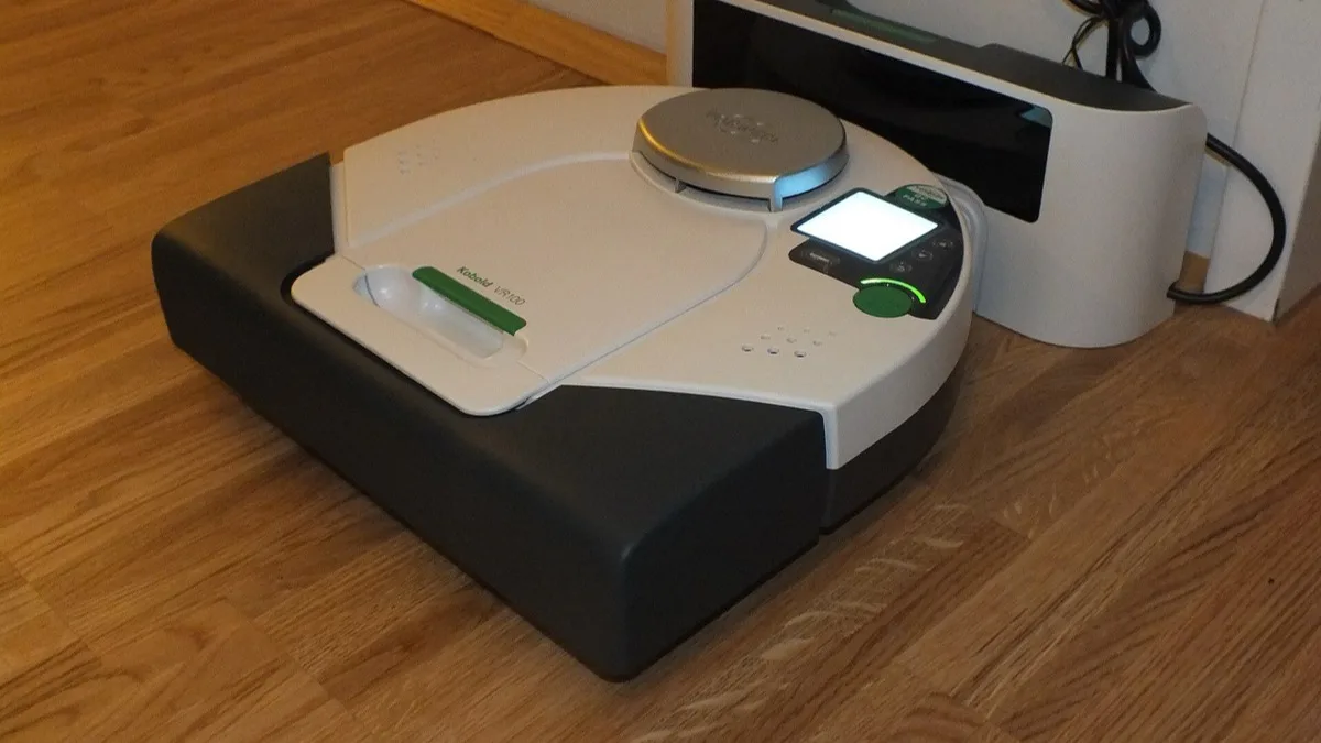 A white and black robot vacuum cleaner parked on its charging dock against a wooden floor