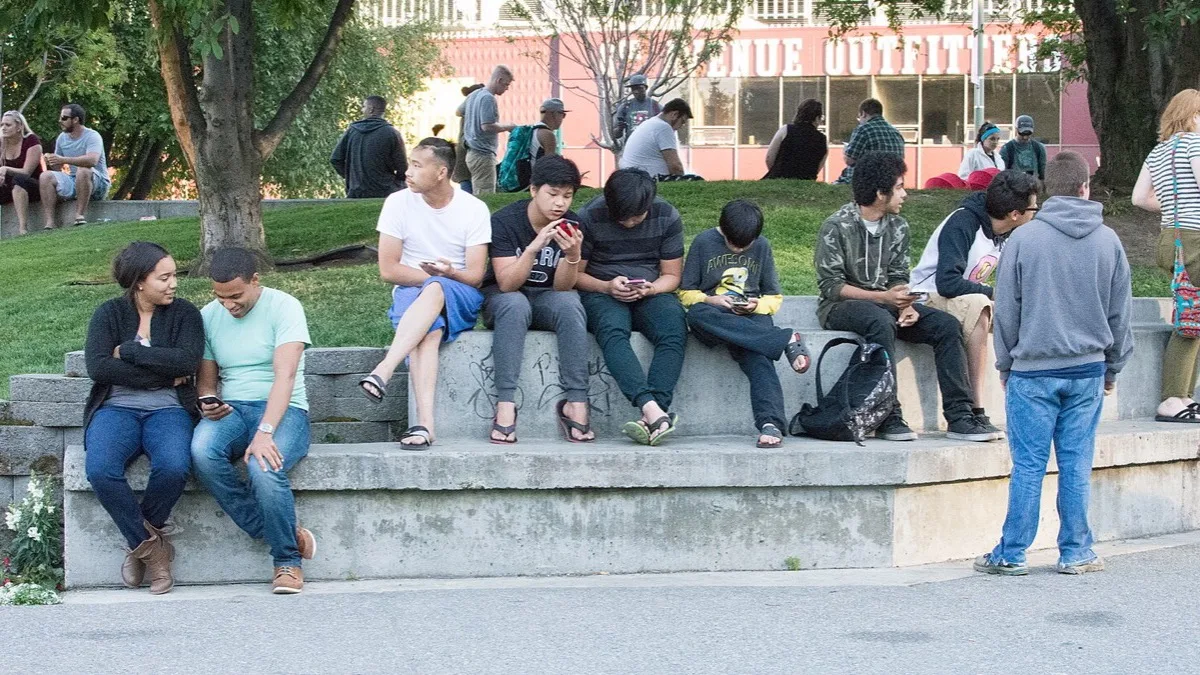 A group of Pokemon GO players sitting on a low concrete wall in a park, each looking at a smartphone