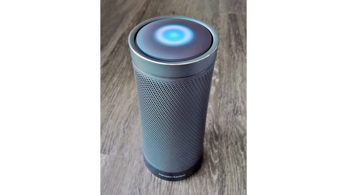 A cylindrical smart speaker with a glowing blue light ring on top, sitting on a wooden floor
