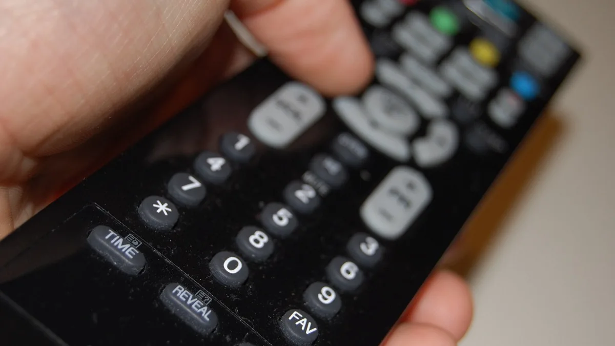 A close-up of a hand holding a black television remote control with numbered buttons