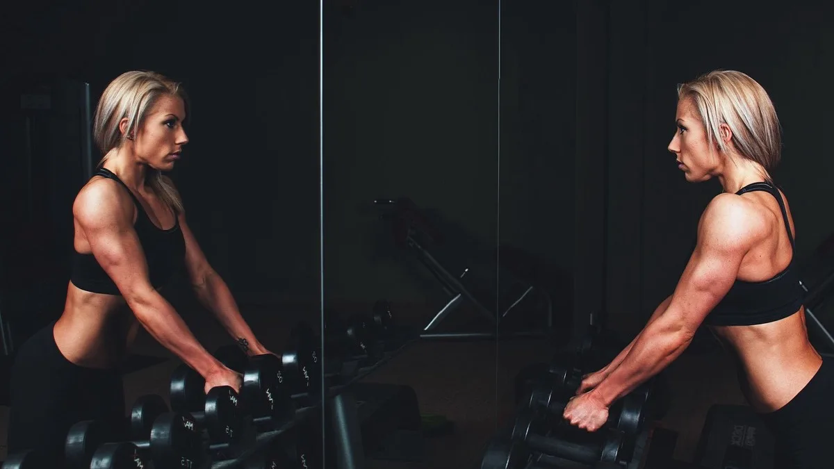 A woman using dumbbells for strength training in a gym, reflected in a wall mirror