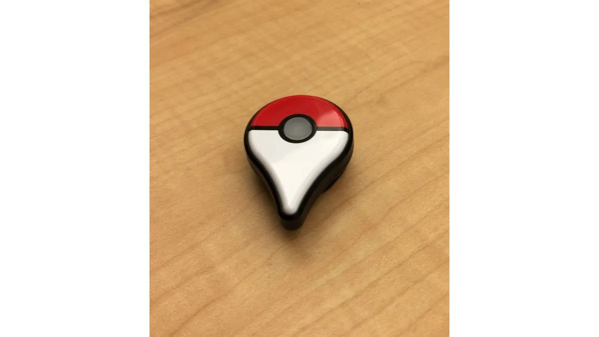 A Pokemon GO Plus wearable accessory, a small red and white Poke Ball-shaped puck, resting on a wooden desk