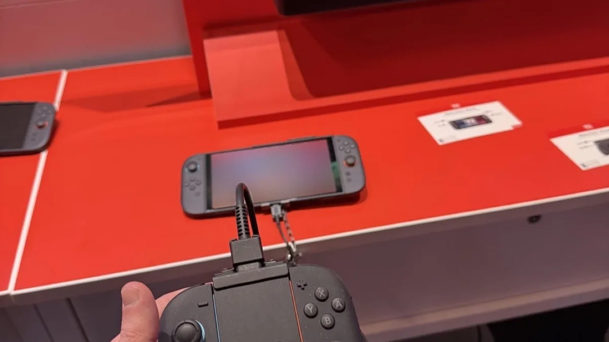 A Nintendo Switch 2 console sitting on a red retail demo counter while a hand holds its Joy-Con 2 controllers in a grip