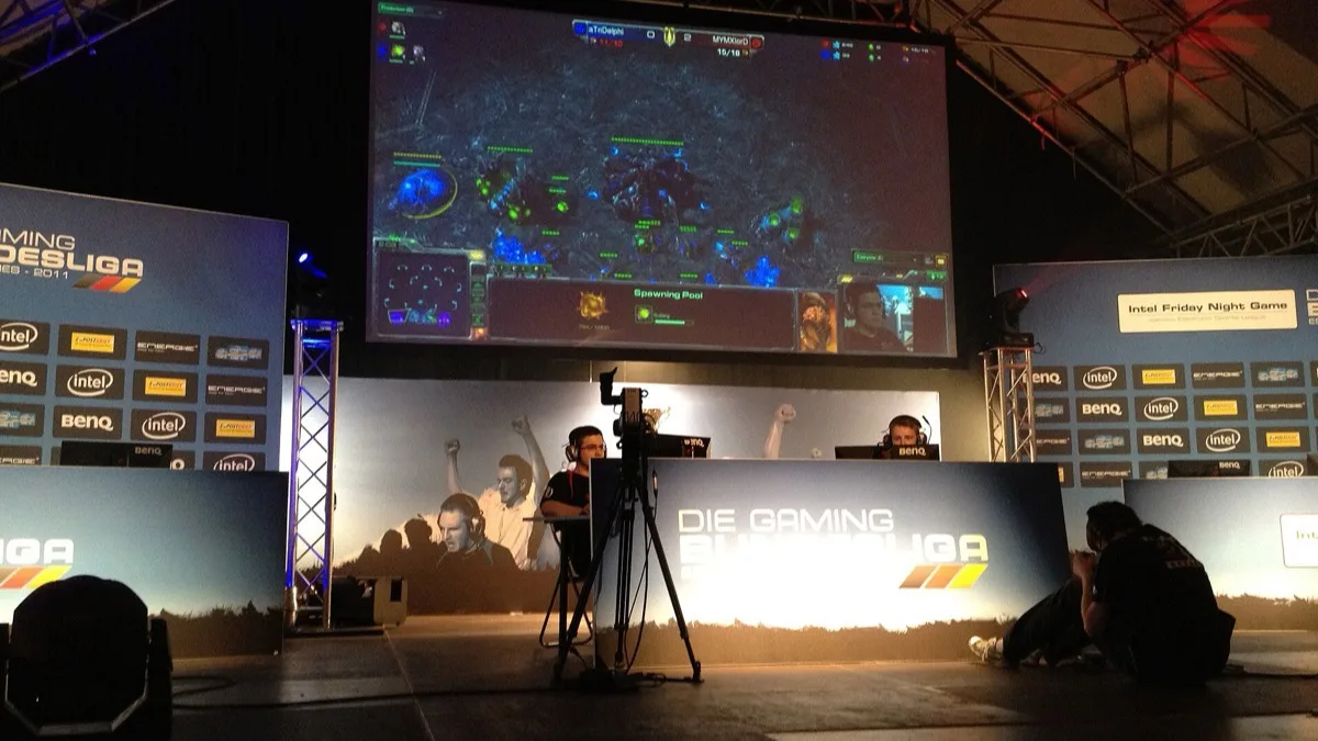 Two players compete on a lit esports tournament stage beneath a large screen showing the live match view