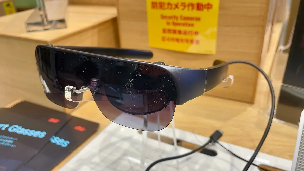 A pair of dark smart glasses with a cable attached, displayed on a stand next to a store sign