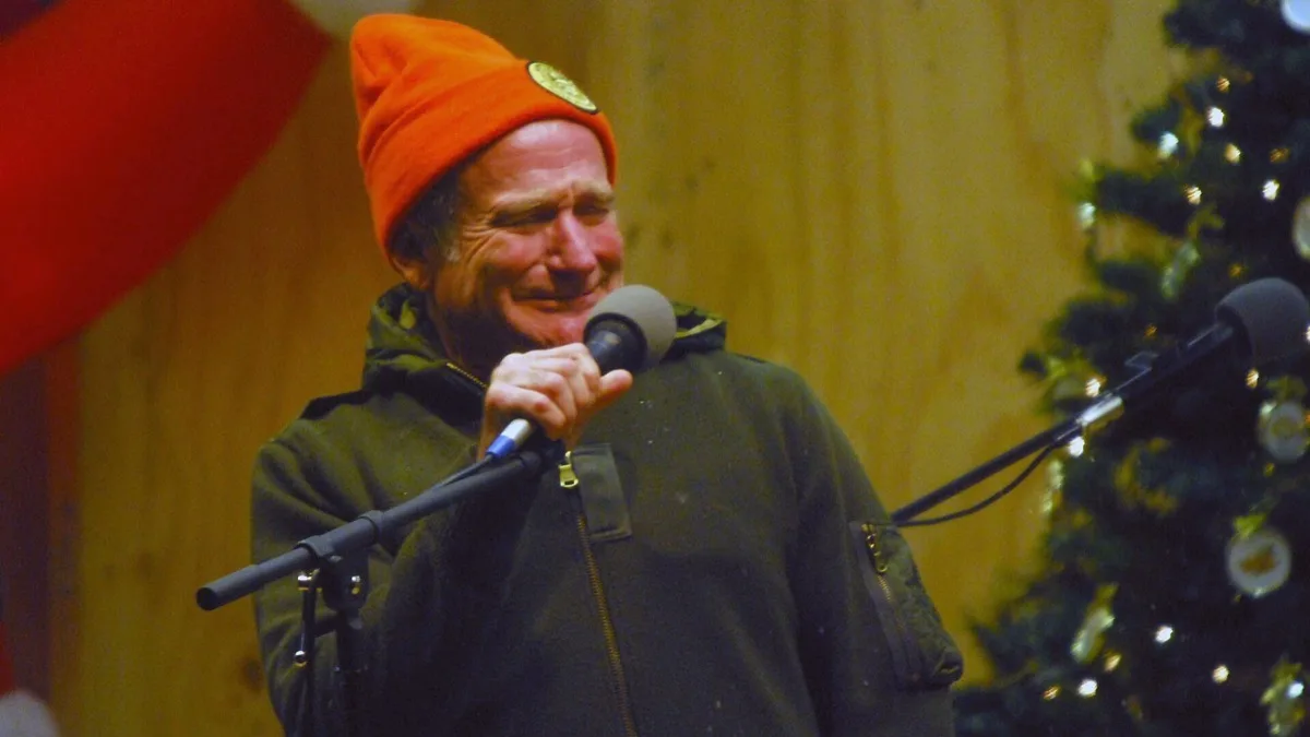 Robin Williams smiling into a microphone in an orange knit cap while performing for troops at Camp Phoenix