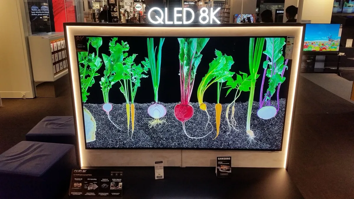 A large Samsung QLED 8K television on display in a store, showing a high-contrast image of vegetables against a dark background