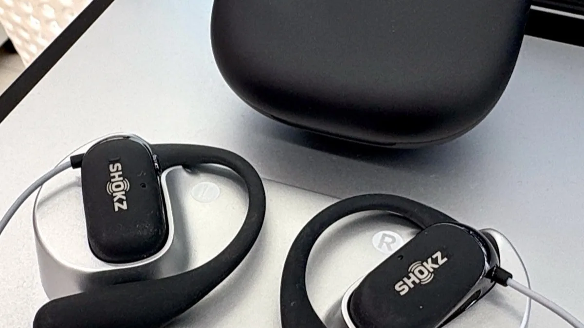 A pair of Shokz open-ear hook earbuds and their charging case resting on a laptop