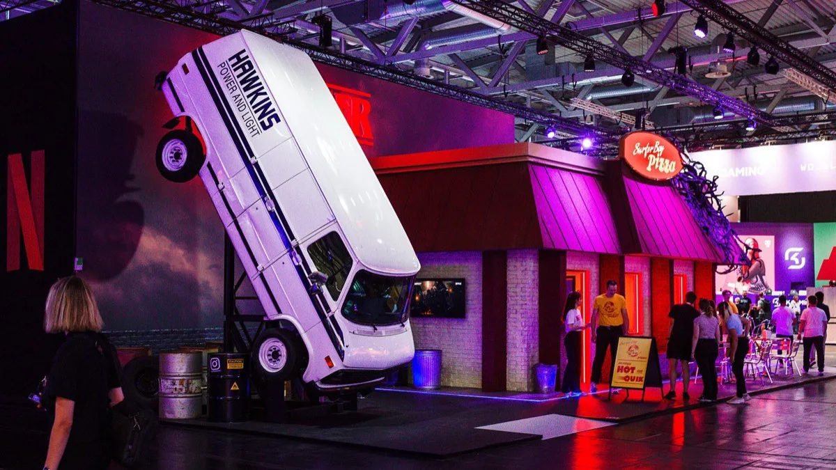 A Stranger Things convention booth with a Surfer Boy Pizza storefront and a Hawkins Power and Light van tipped onto its nose