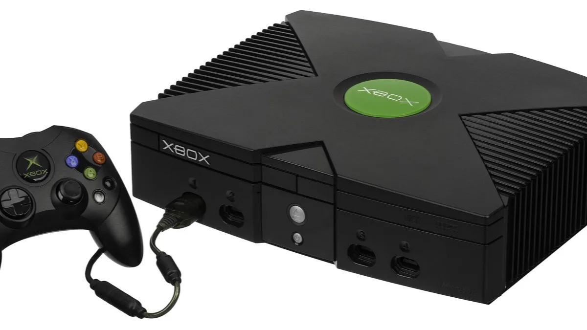 The original black Xbox console from 2001 with its green jewel logo on top, shown beside a black Xbox controller