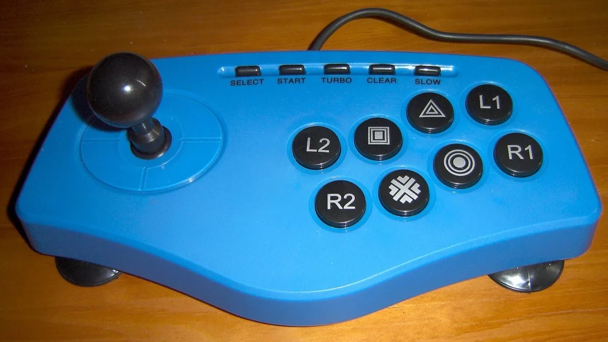 A blue arcade fight stick with a ball-top joystick and eight PlayStation-style face buttons sitting on a wooden desk