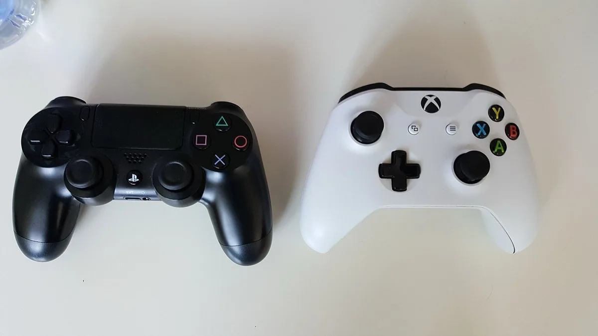 A black PlayStation 4 DualShock 4 controller beside a white Xbox One wireless controller on a table
