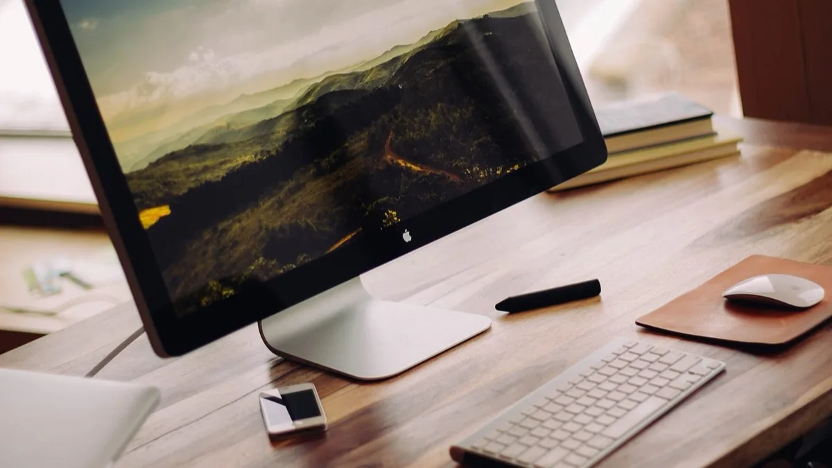 An Apple desktop computer with a wireless keyboard and mouse on a wooden desk beside a stack of books