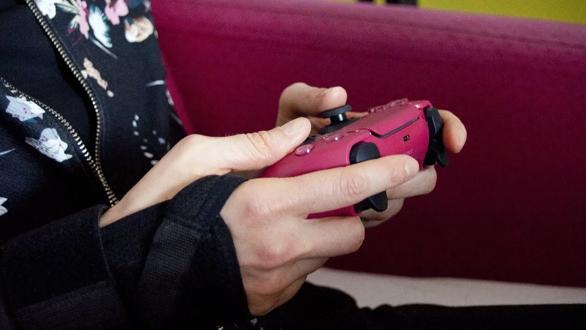 A person wearing a fabric wrist support holds a magenta PlayStation 5 DualSense controller while sitting on a sofa