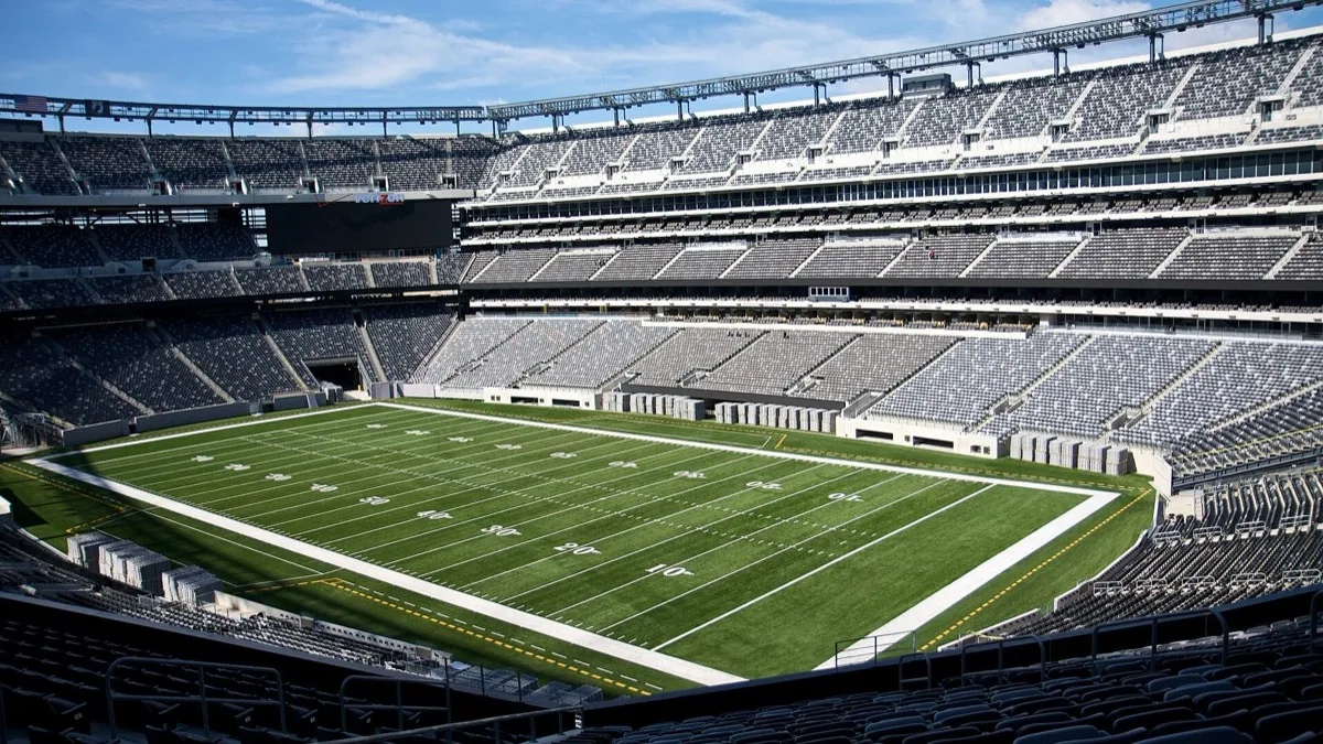 An empty MetLife Stadium seen from a mezzanine corner, with the green field and grey seating bowl under a blue sky