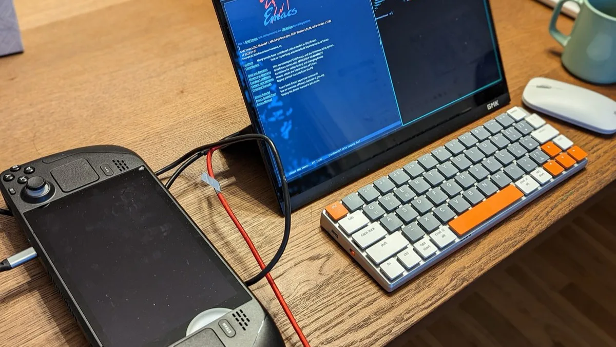 A Steam Deck sitting on a wooden desk, connected by cable to an external monitor running a Linux desktop, with a compact keyboard and mouse beside it