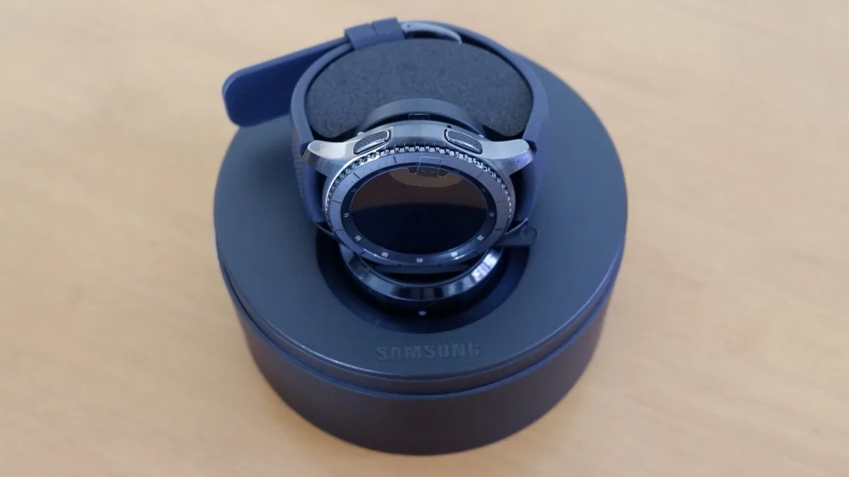 A Samsung Galaxy Watch resting in its circular charging cradle on a wooden desk
