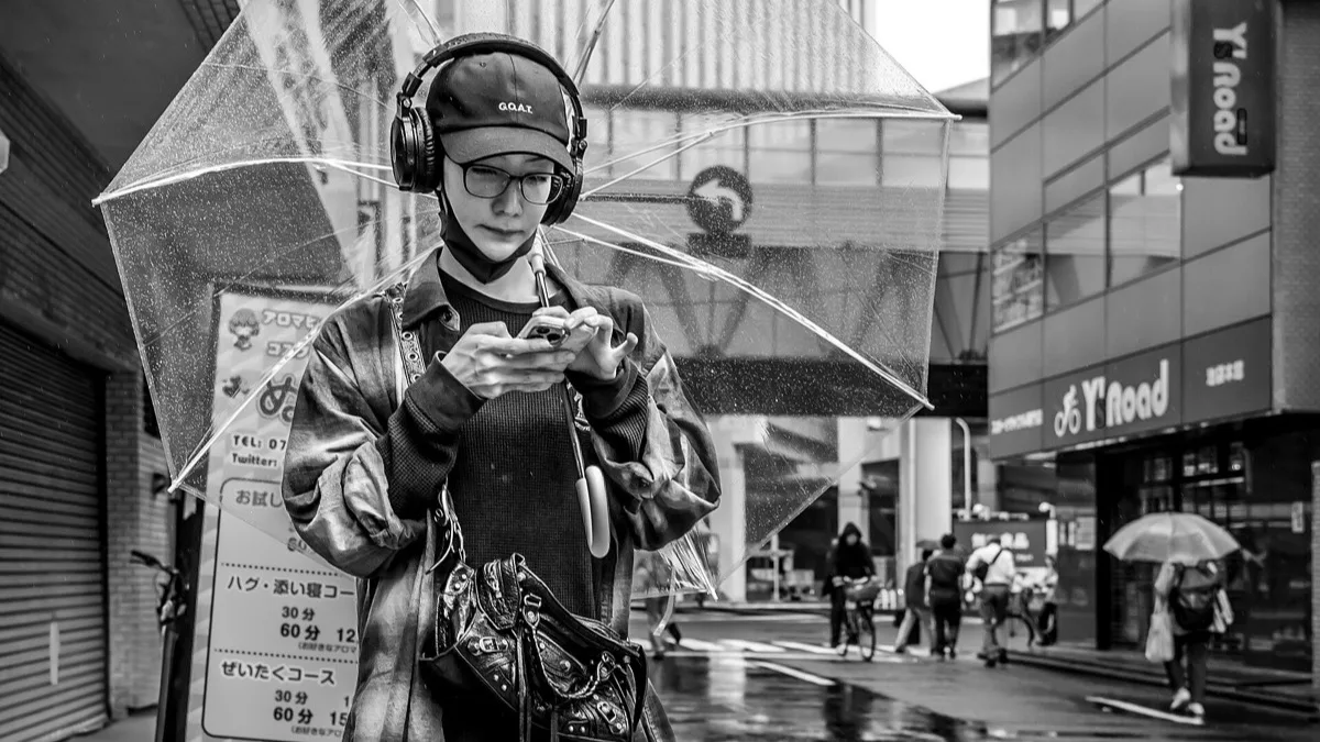 A person in headphones checking a smartphone under a clear umbrella on a rainy street in Japan