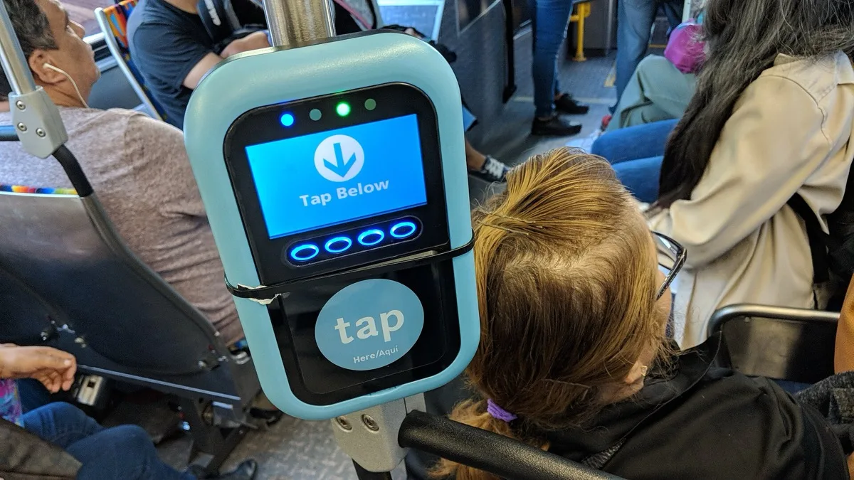 A contactless fare validator on a public bus displaying a Tap Below prompt above a tap here reader pad