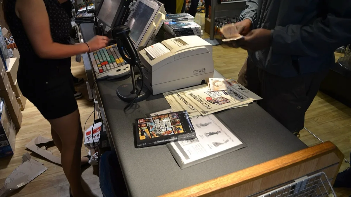 A boxed copy of Grand Theft Auto V sitting on a game shop counter beside a till and a barcode scanner at a midnight launch