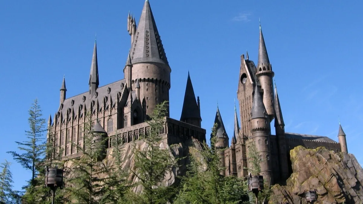 A castle with stone towers and pointed slate spires rising above pine trees against a clear blue sky