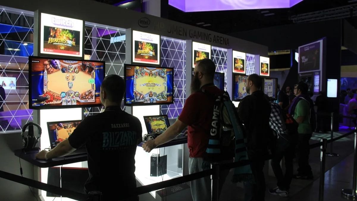 Players standing at Hearthstone match stations in a convention gaming arena, with the game's board shown on several monitors