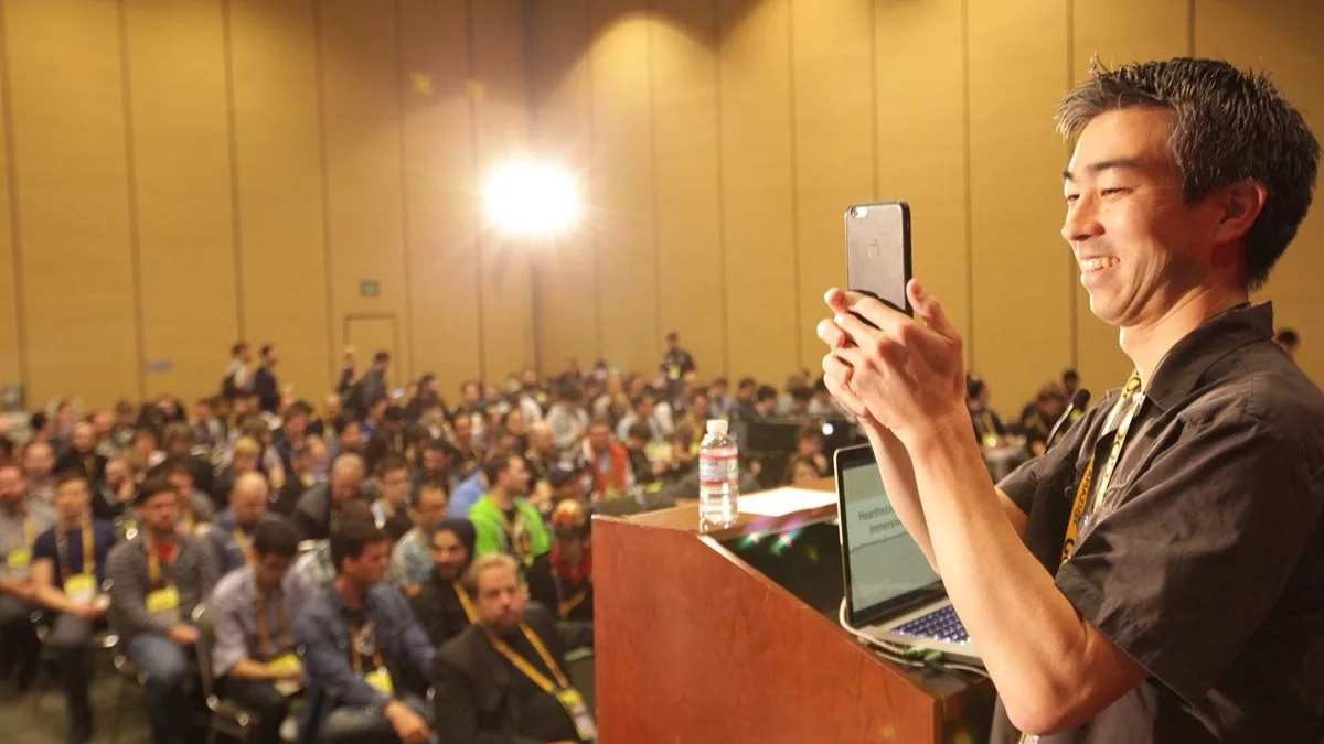 A Blizzard designer at a lectern photographing a packed conference hall with his phone before a Hearthstone talk