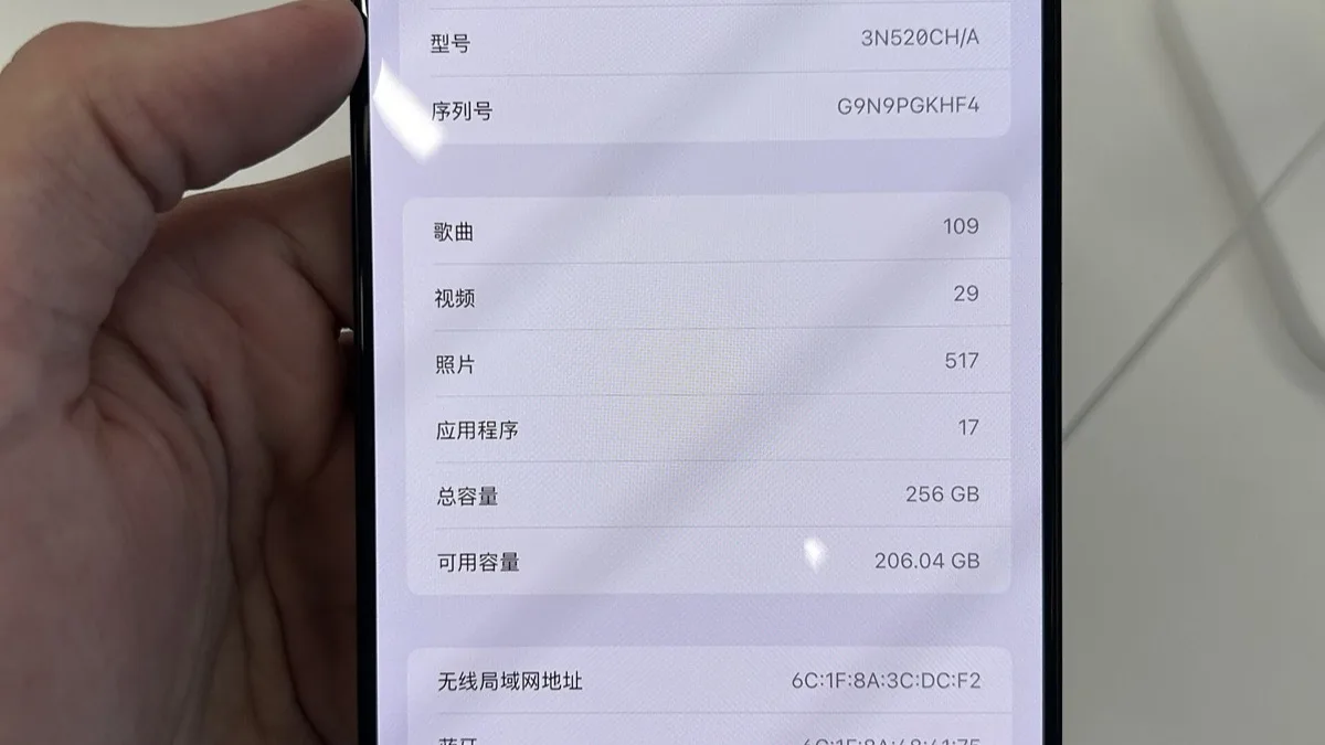 An iPhone held in one hand showing the Settings About screen with model, serial number and storage details