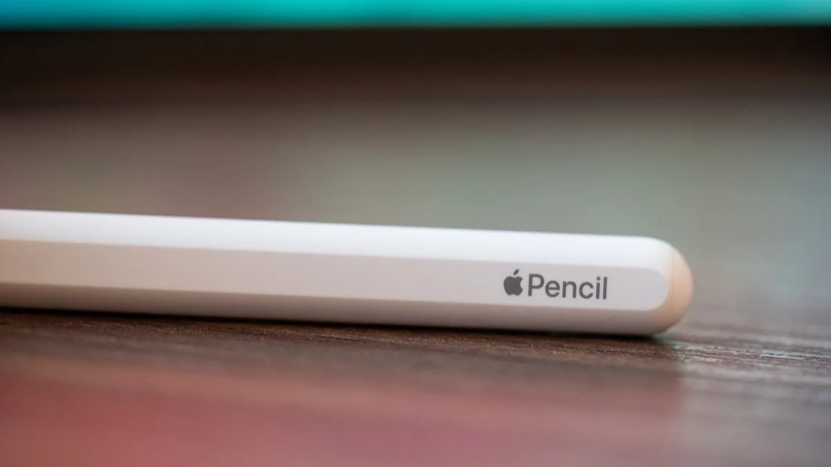 A white Apple Pencil resting on a wooden desk beside a tablet, with the Apple Pencil logo visible on the barrel