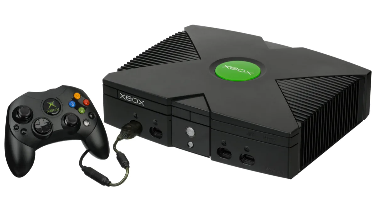 The original black Xbox console from 2001 with its green jewel logo, pictured beside a wired Controller S