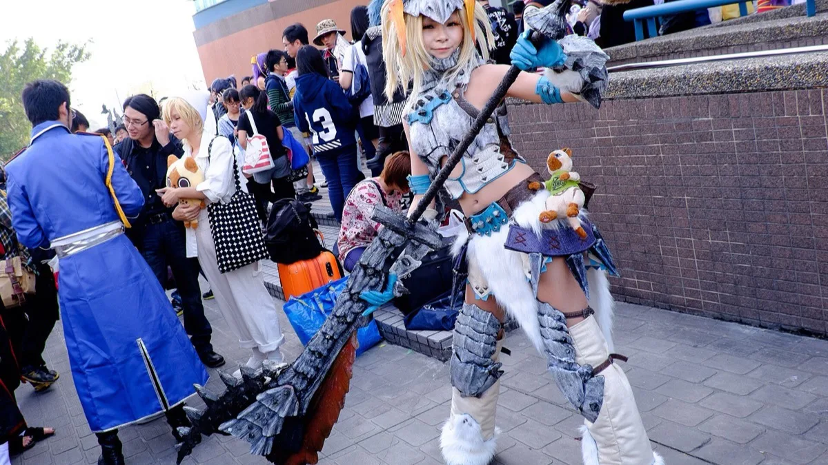 A cosplayer in white fur Monster Hunter armour holding a large jagged great sword, with a Palico plush tied to her belt
