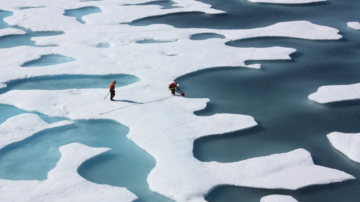Two people in red survival gear cross Arctic sea ice broken up by bright blue meltwater ponds