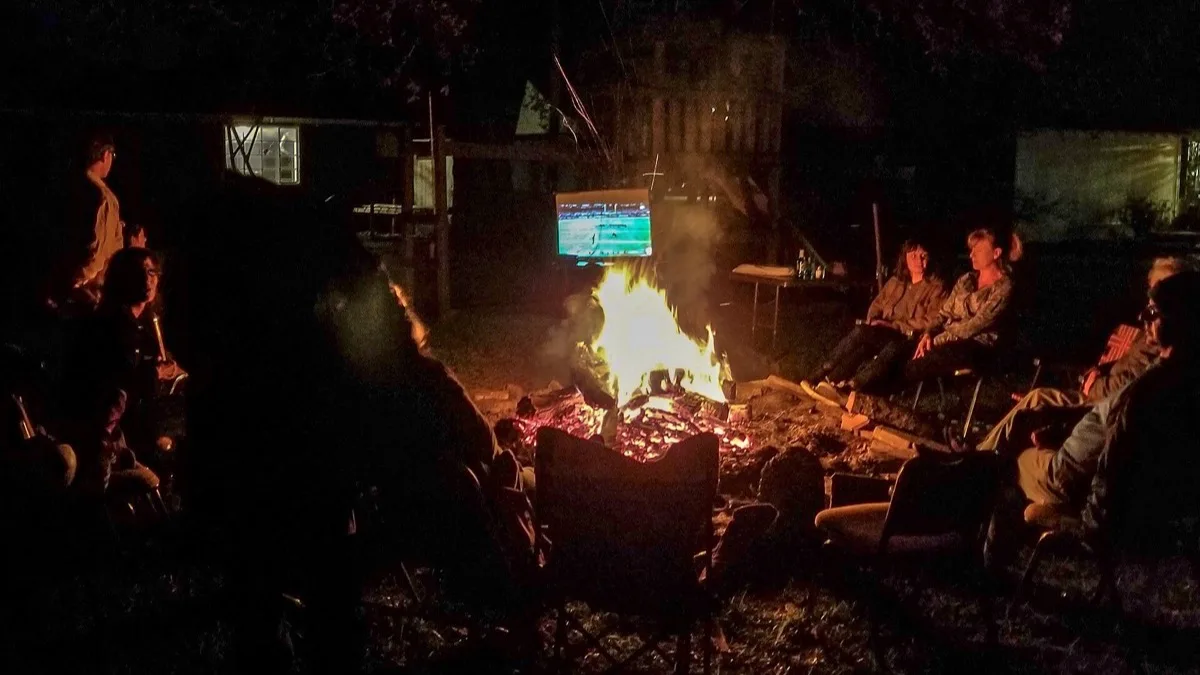 People sitting around a backyard fire at night watching a football game on a television screen