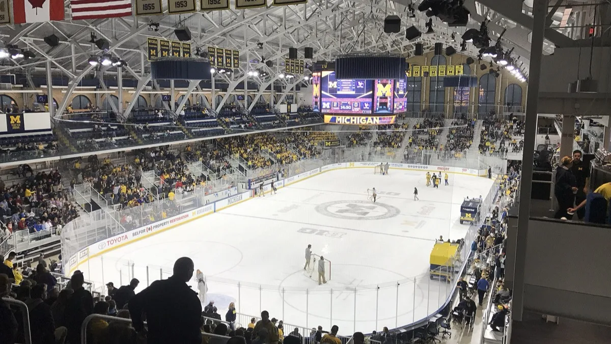 A college ice hockey arena during an intermission, with the rink empty and crowds filling the stands beneath the scoreboard