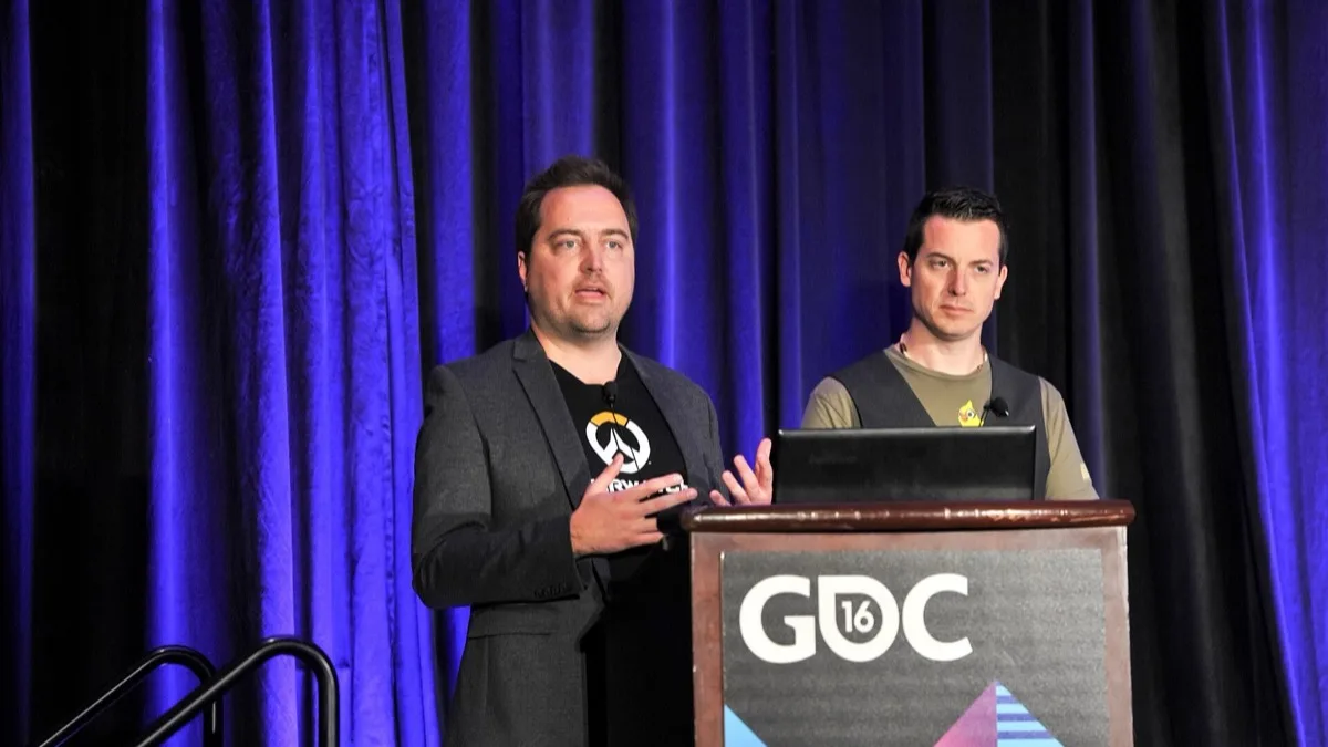 Two Overwatch developers presenting on stage behind a GDC 2016 podium, one wearing an Overwatch logo T-shirt