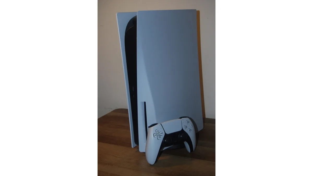 A white PlayStation 5 disc console standing upright with a white DualSense controller resting on the wooden surface in front of it