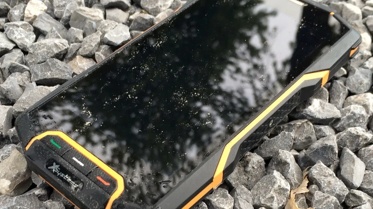 Black and orange rugged waterproof smartphone lying face-up on wet gravel